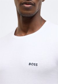 Bílé bavlněné tričko s kulatým výstřihem a krátkými rukávy, s černě vyšívaným logem "BOSS" na levé straně hrudi. Hladká textura.