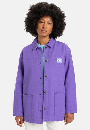 Chaqueta de algodón morado con un collar, cierre delantero con botones, dos bolsillos y un logo impreso en el pecho. Color sólido con una textura suave.