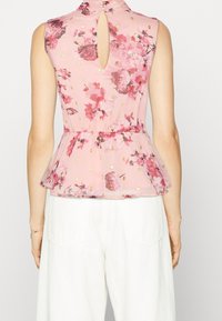 Květinový peplum top v růžové průsvitné tkanině s klíčovou dírkou na zadní straně. Obsahuje zlaté akcenty a volánkový lem. Doplněno o bílé kalhoty s širokými nohavicemi.