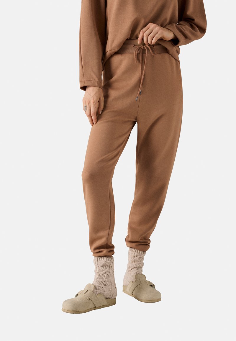 Pantalones jogger de tela suave y marrón, con cintura elástica y cordón ajustable. Combinados con zuecos color crema y calcetines de punto rosa claro.