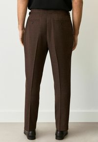 Pantalons marron taillés avec une finition texturée, dotés de deux poches arrière et d'un pli le long de la jambe, associés à des chaussures noires.