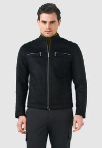 Giacca in suede nera con colletto alto, doppia zip frontale e quattro tasche con zip. Le maniche trapuntate testurizzate aggiungono dettaglio.