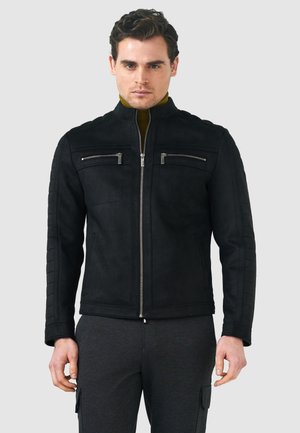 Giacca in suede nera con colletto alto, doppia zip frontale e quattro tasche con zip. Le maniche trapuntate testurizzate aggiungono dettaglio.
