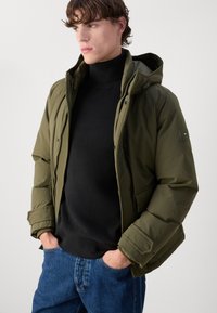 Olivengrøn pufferjakke med hætte, lynlåslukning foran og sidelommer, parret med en sort tekstureret sweater og blå jeans.