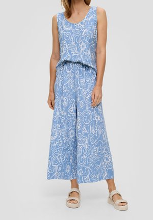 Kobieta ubrana w niebieski kombinezon bez rękawów z białymi kwiatowymi wzorami i szerokimi nogawkami, w połączeniu z beżowymi plecionymi sandałami.