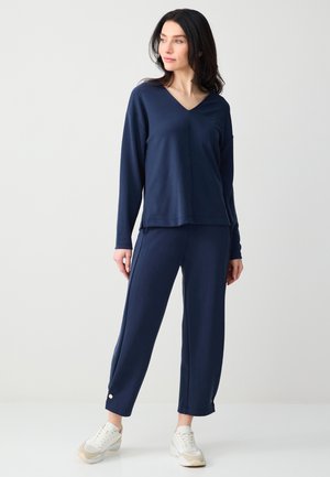 Kobieta stojąca, ubrana w granatową bluzkę z długim rękawem i dopasowane spodnie oraz białe trampki, patrząca w bok na tle jednolitego tła.