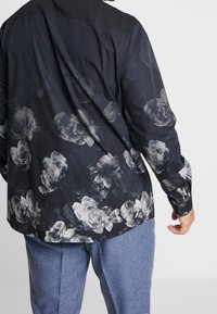 Homme portant une chemise noire à manches longues avec un motif floral gris et un pantalon bleu, vu de dos sur un fond blanc uni.