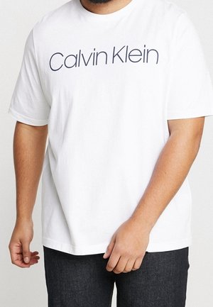 Mann trägt ein weißes Calvin Klein T-Shirt und dunkle Jeans, steht mit entspannt an den Seiten hängenden Händen.