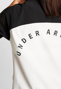 Chaqueta en blanco y negro que presenta una sección superior completamente negra y una sección inferior blanca con el texto impreso "UNDER ARMOUR" en negro.