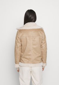 Beige mockajacka med shearlingkrage och manschetter, med vit sömnad och rak design, bärs med ljusa byxor.