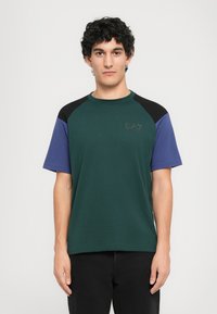 Uomo con capelli ricci neri che indossa una t-shirt a maniche corte verde, nera e blu e pantaloni neri, in piedi su uno sfondo bianco semplice.