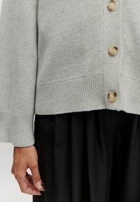 Hellgraue Strickjacke mit geripptem Saum und Bündchen, versehen mit großen cremefarbenen Knöpfen entlang der Vorderseite, getragen über einem schwarzen Rock.