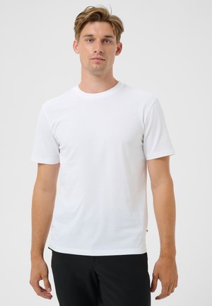 Joven con cabello castaño claro, vistiendo una camiseta blanca sencilla de manga corta y pantalones negros, de pie frente a un fondo blanco.