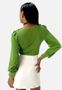 Top verde a maniche lunghe con spalle arricciate e design aderente, abbinato a una mini gonna bianca. La texture del tessuto è liscia.