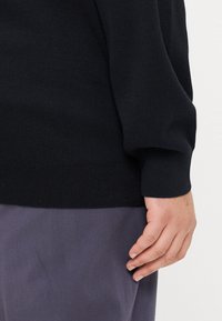 Pulover negru cu tiv și manșete striate, textură moale, purtat peste pantaloni purpurii, arătând o parte dintr-o mână sprijinită de îmbrăcăminte.