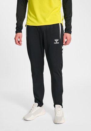 Mężczyzna w czarnych spodniach dresowych Hummel z białymi paskami i logo, białych trampkach oraz żółto-czarnym sportowym topie z długim rękawem.