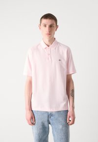 Polo rose clair avec col à deux boutons, manches courtes et petit logo sur la poitrine gauche ; associé à un jean en denim bleu clair.