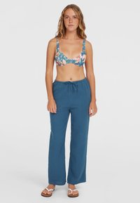 Floral-patterned bralette in shades of blue and pink with adjustable straps, paired with loose, dark blue, breathable pants and white sandals.