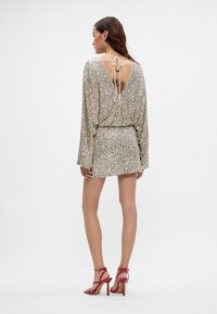 Šimerasta zlata obleka s palčkami in globokim V-izrezom na hrbtni strani, zategnjena v pasu. V kombinaciji z rdečimi sandali na visokih petah.