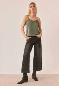 Top verde sin mangas con ribete de encaje, combinado con jeans negros de pierna ancha y cintura alta. La modelo lleva botas negras hasta el tobillo y tiene el cabello lacio.