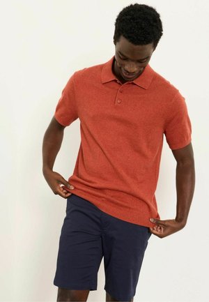 Mann passt Saum eines orangefarbenen Poloshirts mit kurzen Ärmeln an, trägt marineblaue Shorts, steht vor einem schlichten weißen Hintergrund.