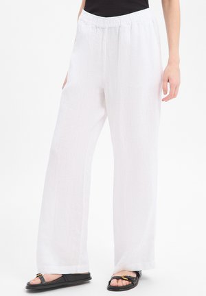 Vrouw draagt witte linnen broek met wijde pijpen en elastische tailleband, met zwarte sandaal met open teen, staand tegen een witte achtergrond.