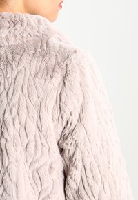 Syntetisk pälsjacka i ljusrosa, med ett texturerat vågmönster och mjuk yta. Kragen är rundad och tyget verkar tjockt och varmt.