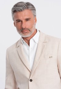 Blazer de lino beige claro con una textura sutil, cierre de botón único y un adorno en el bolsillo del pecho, sobre una camisa blanca.