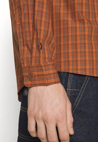 Gros plan sur une main à côté d'un poignet de chemise à carreaux marron avec bouton, associée à un jean en denim foncé avec des coutures jaunes et une petite étiquette.