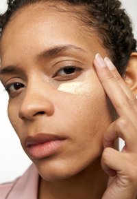 Hautpflegeanwendung mit einem helltonigen Concealer-Patch unter dem Auge. Glatte Hautstruktur, natürlicher Make-up-Look und lockige Haare.