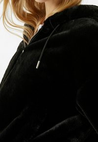 Svart hoodie i konstläder med mjuk textur, med dragkedja och justerbara snörning. Tyget verkar plyscht och varmt.