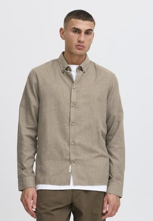 Mand iført en beige langærmet skjorte knappet op over en hvid undertrøje og brune bukser, stående mod en ensartet lys grå baggrund.