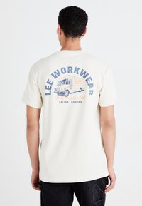 Grietos spalvos medvilninis T-shirt marškinėliai su grafikos spauda ant nugaros, vaizduojančia senovinį sunkvežimį, mėlyną tekstą ir oranžinius akcentus. Trumpomis rankovėmis, laisvo modelio.