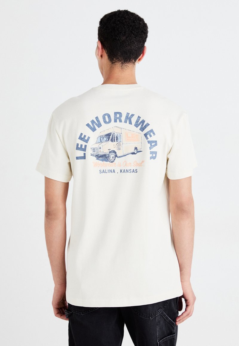 Grietos spalvos medvilninis T-shirt marškinėliai su grafikos spauda ant nugaros, vaizduojančia senovinį sunkvežimį, mėlyną tekstą ir oranžinius akcentus. Trumpomis rankovėmis, laisvo modelio.