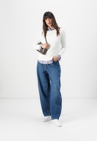 Hvit genser med rund hals, lagdelt over en blå og hvit stripete skjorte, parret med løse blå jeans og hvite joggesko.