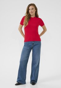 Haut rouge à manches courtes côtelé avec un col rond, associé à un jean large bleu clair et des chaussures noires, sur fond uni.