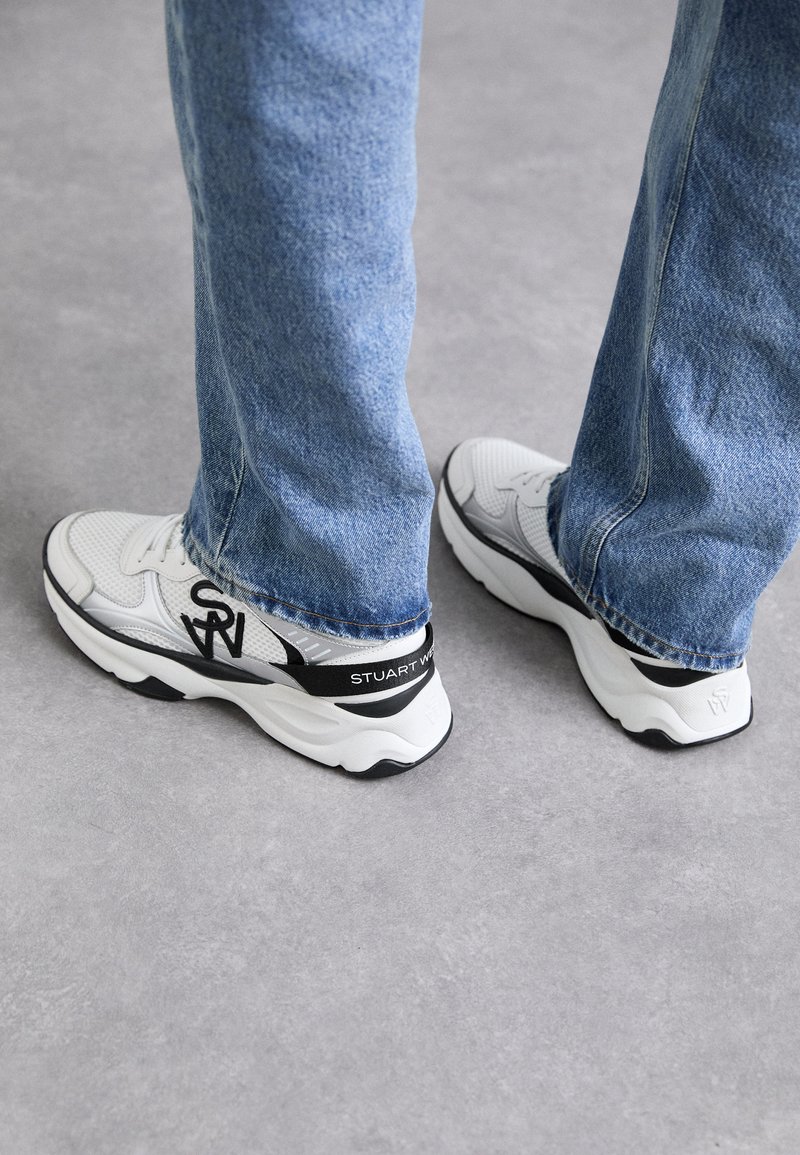 White and gray athletic shoes with a textured mesh upper, black accents, and a prominent logo on the side and heel, paired with blue jeans.