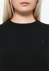 Crna pletena majica s okruglim izrezom i rebrastom teksturom, s malim reljefnim logotipom na prsima. Mekani materijal.