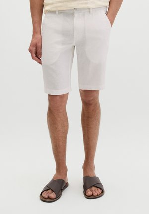 Męskie nogi ubrane w białe szorty do kolan oraz ciemnobrązowe sandały z krzyżującymi się paskami, stojące na jednolitym tle.