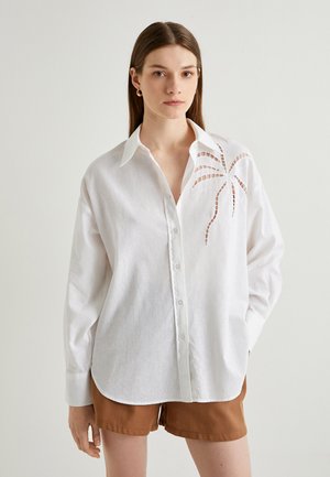 Camisa de lino blanca con botones en el frente, cuello puntiagudo y diseño de palmera recortada en el hombro izquierdo. Mangas largas, corte relajado.