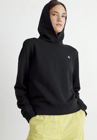 Czarna bluza z kapturem wykonana z gładkiego materiału, z małym logo na piersi i ściągaczami przy rękawach, zestawiona z żółtymi spodniami.