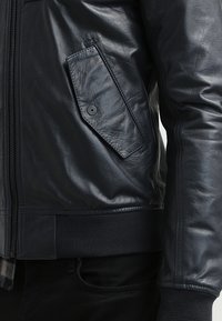 Mörkblå läderjacka med dragkedja, sidoficka med knapp, ribbade ärmslut och en slät, blank yta.