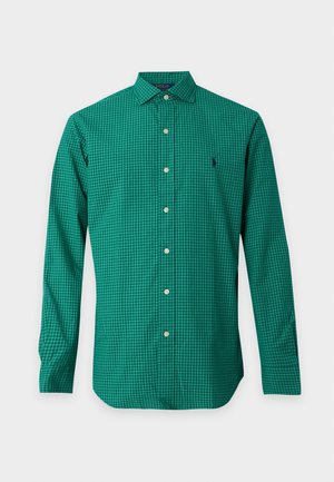 Homme portant des lunettes de soleil noires, une chemise à carreaux verte à manches longues, un pantalon jaune moutarde et des baskets blanches, debout avec une main dans la poche.
