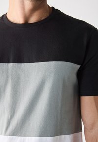 Tricou din bumbac negru, gri și alb, cu mâneci scurte. Prezintă un design de tip color-block orizontal în secțiuni tonale solide. Textură moale.