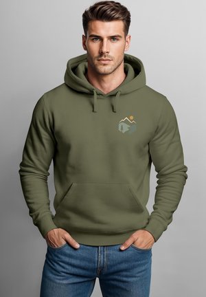 Mann mit kurzen braunen Haaren, der einen olivgrünen Hoodie mit Berg- und Baum-Design trägt und mit den Händen in den Taschen einer blauen Jeans steht.