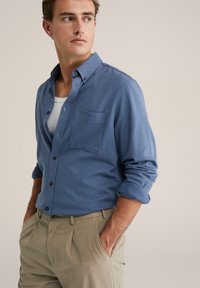 Kék gombos ing mellzsebbel, texturált anyagból és felhajtott ujjal, bézs nadrággal párosítva. Semleges háttér.