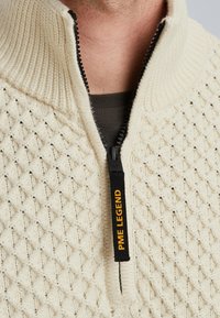 Beige gebreide trui met een gestructureerd ruitpatroon, voorzien van een hoge ritssluiting, contrasterende zwarte rits en een label met "PME LEGEND" in het geel.
