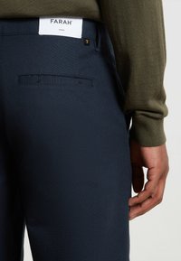 Marineblauwe broek met een gladde textuur, voorzien van een achterzak en een wit merklabel op de tailleband. Olijfgroene trui-mouw zichtbaar.