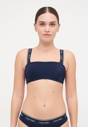 Navy blue bandeau bikini top with wide straps featuring white "Calvin Klein" branding. Smooth material with a fitted silhouette.