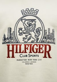 Κεντητός λέοντας που κρατάει σπαθί μέσα σε κύκλο, φόντο με ορίζοντα πόλης, κείμενο κάτω που αναφέρει «HILFIGER Club Sports» και «Manhattan New York City».